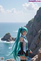 A woman with long blue hair standing on a cliff by the ocean.